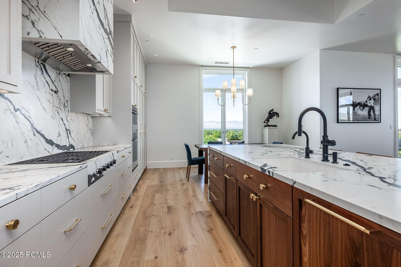 This is a bright and modern kitchen featuring white and wood cabinetry with gold hardware, marble countertops and backsplash, and stainless steel appliances. The kitchen island provides ample counter space and a sink, while a dining area is visible in the background. The overall impression is one of luxury and sophistication.