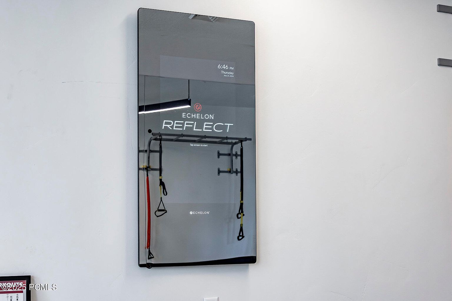The image showcases an Echelon Reflect fitness mirror mounted on a white wall, reflecting a home gym setup with suspension training equipment. The mirror displays the time and date, adding a modern touch to the workout space. The overall impression is a clean and technologically advanced home fitness area.