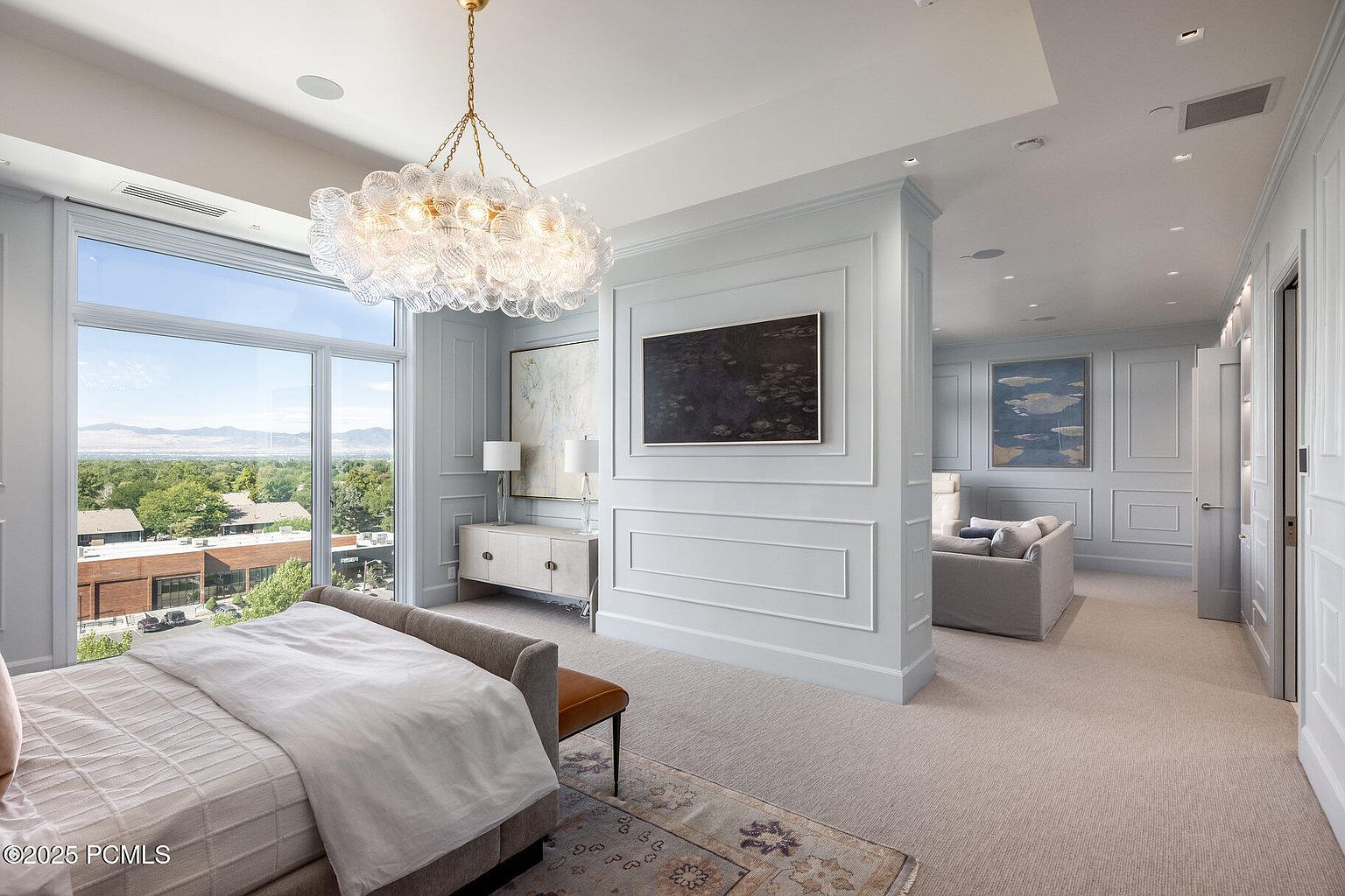 This is a luxurious primary bedroom featuring a large window with scenic views, a statement chandelier, and elegant wall paneling. The room includes a bed with neutral linens, a console table with lamps, and artwork, leading into a sitting area. The overall impression is one of sophistication and comfort.