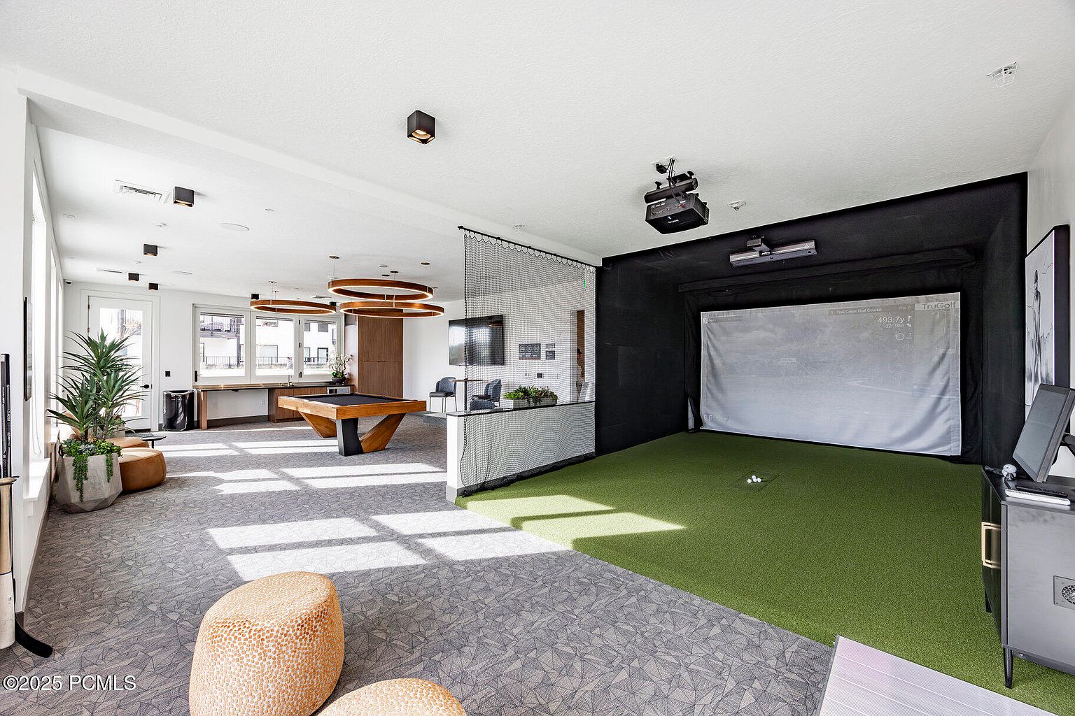 This interior shot showcases a community amenity space, featuring a golf simulator and a billiards table. The room is well-lit with modern lighting fixtures and has a contemporary design with a neutral color palette and pops of green from the golf simulator area and plants. The space appears to be designed for recreation and entertainment.