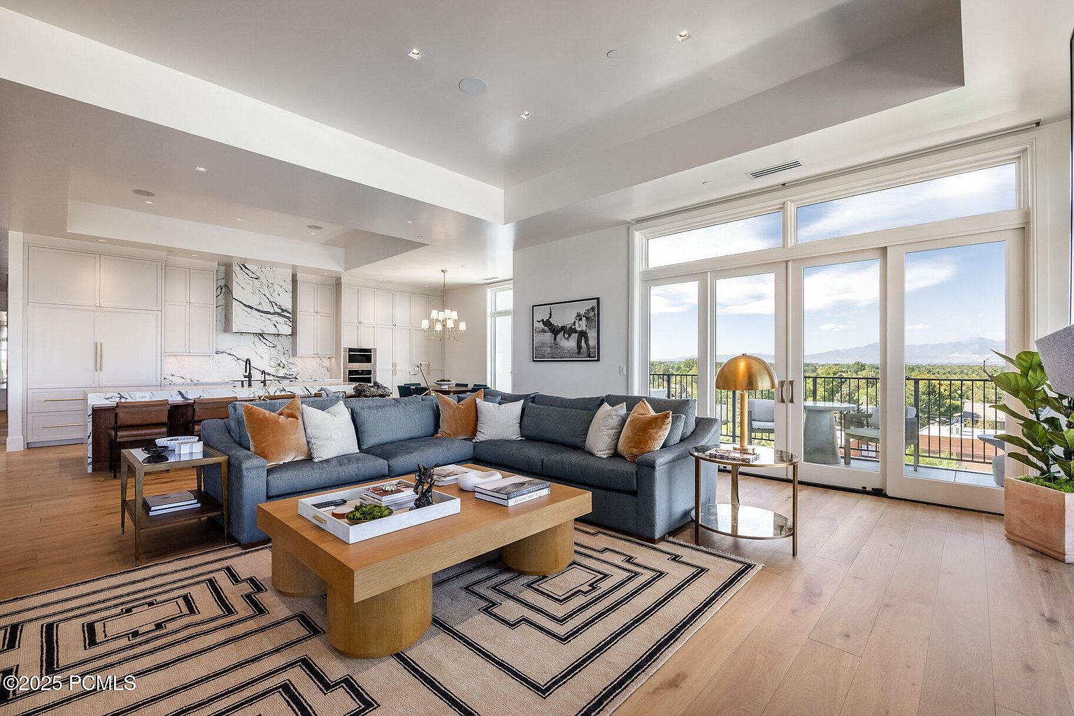 This is a spacious and bright living room that seamlessly blends with the kitchen area. The room features a large sectional sofa with decorative pillows, a modern coffee table, and a patterned area rug. Large windows offer a stunning view of the outdoors, and the overall design is contemporary and inviting.