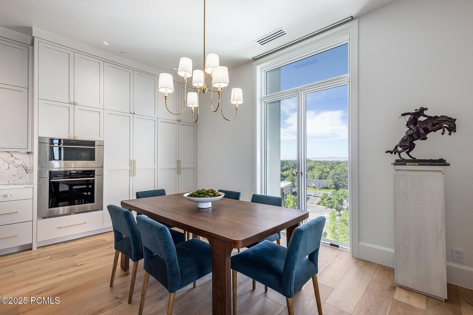This dining room features a wooden table with six blue velvet chairs, illuminated by a modern chandelier. Built-in white cabinets and a stainless steel oven are visible along one wall, while a large window offers a view of the outdoors. A sculpture on a pedestal adds an artistic touch to the space.