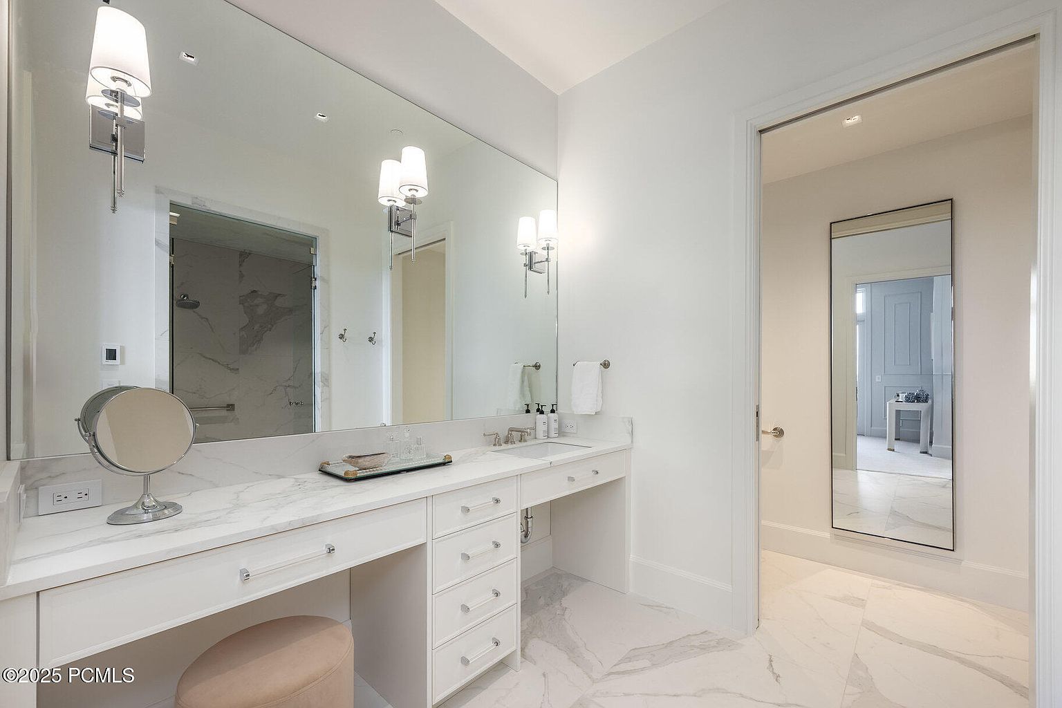 This is a bright and elegant primary bathroom featuring a large vanity with a marble countertop, ample storage drawers, and a makeup area with a stool. A large mirror spans the length of the vanity, reflecting the room and enhancing the sense of space. The flooring is marble, and a doorway leads to another room, possibly a closet or dressing area.