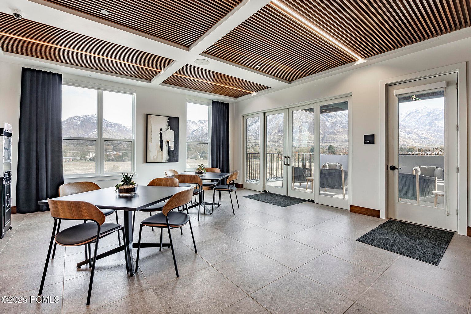This interior shot showcases a modern dining area with sleek, dark-topped tables and stylish chairs featuring wood and black accents. The room is well-lit, with natural light streaming through large windows that offer scenic mountain views. A unique slatted ceiling design adds architectural interest, and the overall aesthetic is clean and contemporary, perfect for a clubhouse or community space.