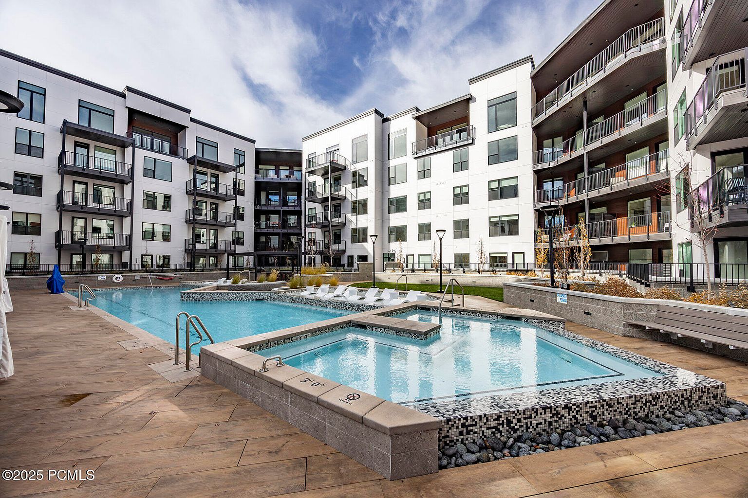 This image showcases a luxurious outdoor pool and spa area within a modern apartment complex. The pool is surrounded by a wooden deck and features a separate spa area with mosaic tile detailing. The contemporary architecture of the buildings provides a stylish backdrop, enhancing the appeal of this community amenity.