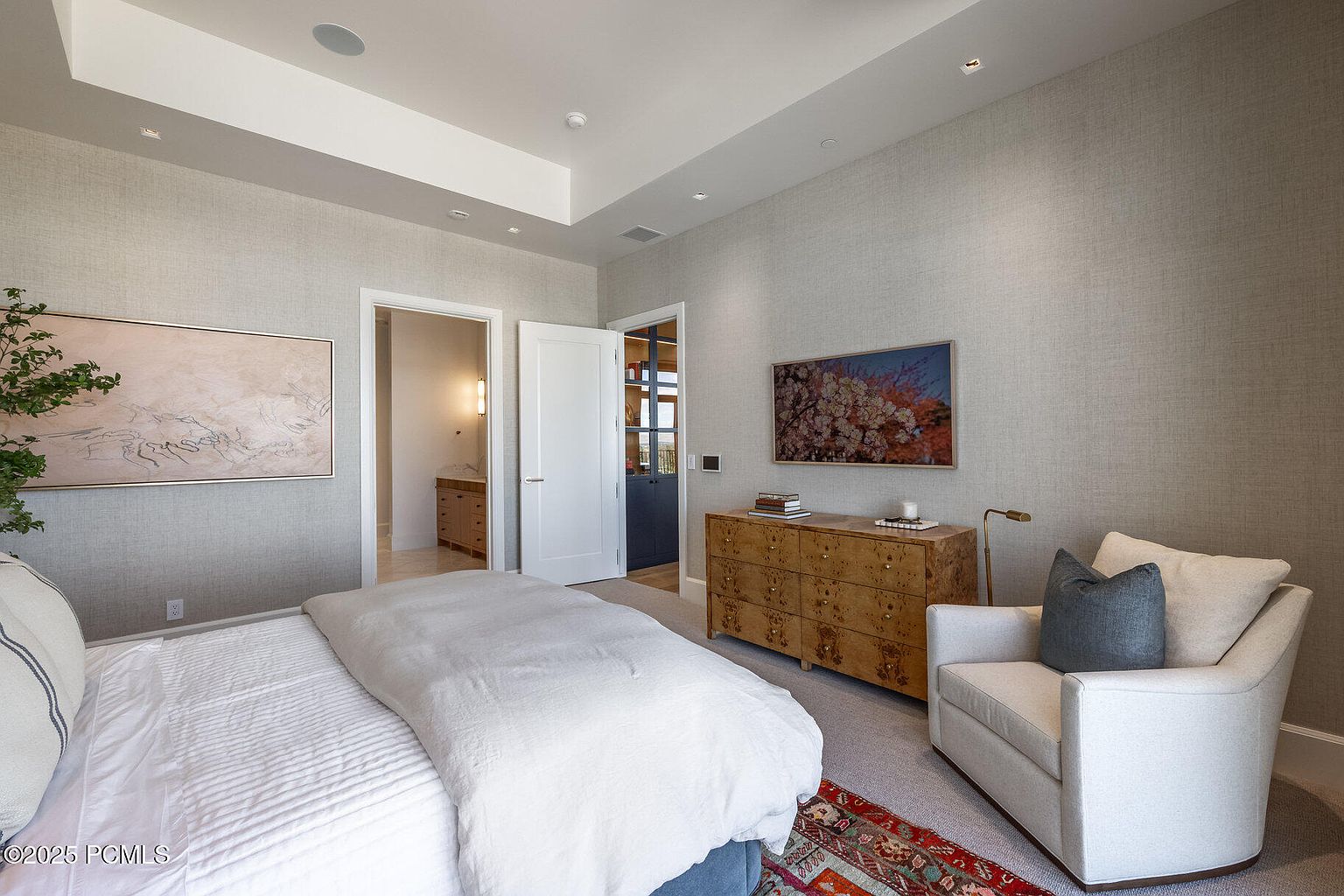This is a primary bedroom featuring a king-size bed with white linens, a comfortable armchair, and a wooden dresser with a television mounted above. The walls are covered in a textured wallpaper, and there are two doorways leading to other rooms. The overall impression is one of comfort and understated elegance.