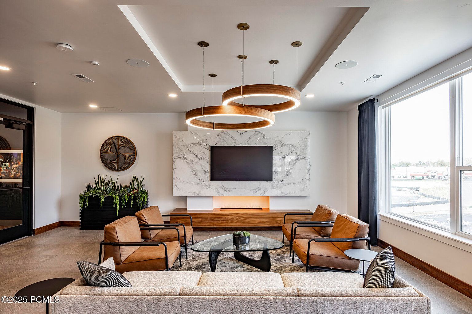 This is a modern living room featuring a neutral color palette with pops of brown from the leather chairs. The focal point is a marble-look wall with a large television and a linear fireplace below. The room is well-lit with natural light from a large window and unique circular pendant lighting, creating a welcoming and stylish atmosphere.