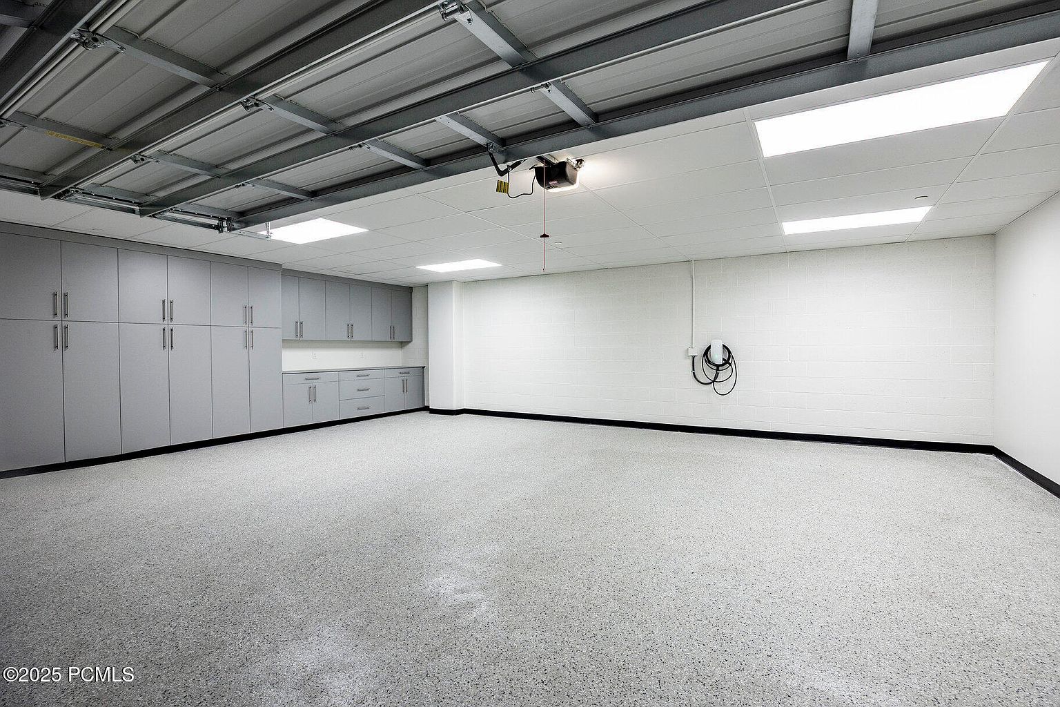 This is an interior shot of a spacious and well-organized garage. The garage features ample storage with gray cabinets along one wall, a clean epoxy floor, and a white ceiling with recessed lighting. The walls are painted white, and a black trim runs along the base, giving the space a modern and functional feel.