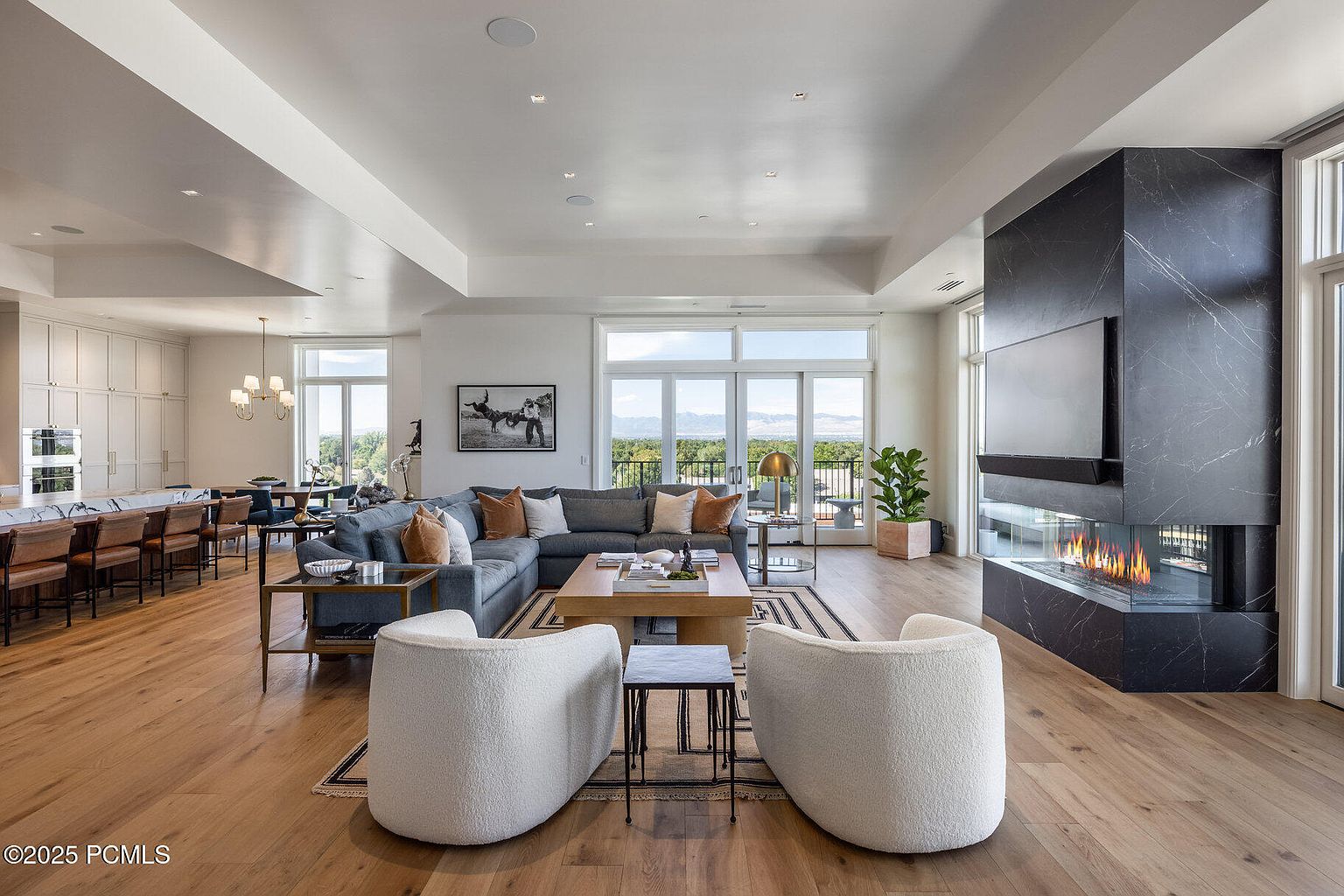 This is a spacious and luxurious living room featuring hardwood floors, a modern fireplace with a television above, and large windows offering scenic views. The room is furnished with a comfortable sectional sofa, two plush armchairs, and stylish coffee tables, creating an inviting and sophisticated atmosphere. The open-concept design seamlessly connects the living area to the dining and kitchen spaces.