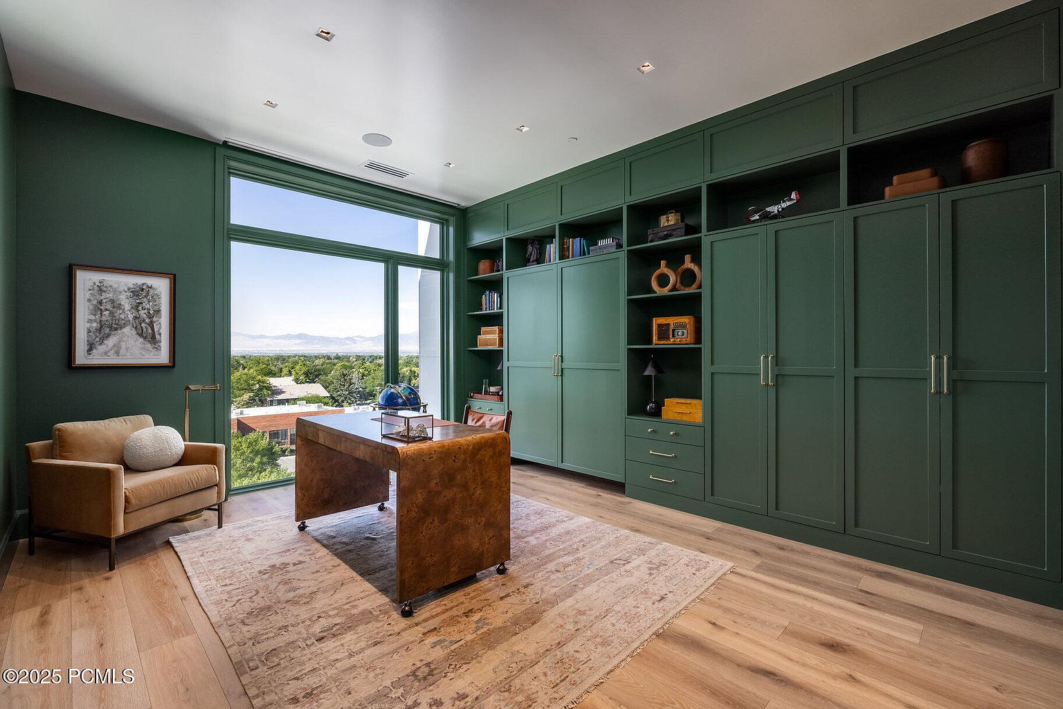 This is an interior shot of a sophisticated home office featuring a custom built-in shelving unit and cabinetry in a deep green hue. A unique, leather-wrapped desk sits in the center of the room, complemented by a comfortable armchair and a patterned area rug. Large windows offer a view of the landscape, enhancing the room's natural light and creating a serene workspace.