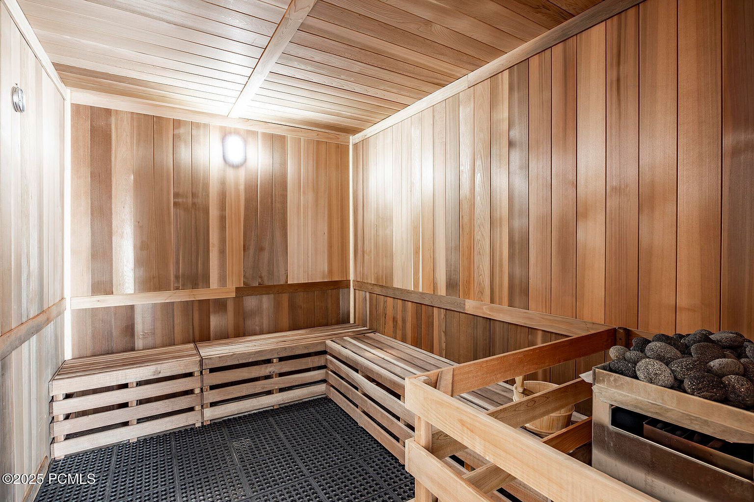This is an interior shot of a sauna, featuring wooden walls, ceiling, and benches. A heater with sauna stones is visible in the corner. The room is well-lit, creating a warm and inviting atmosphere, perfect for relaxation and wellness.