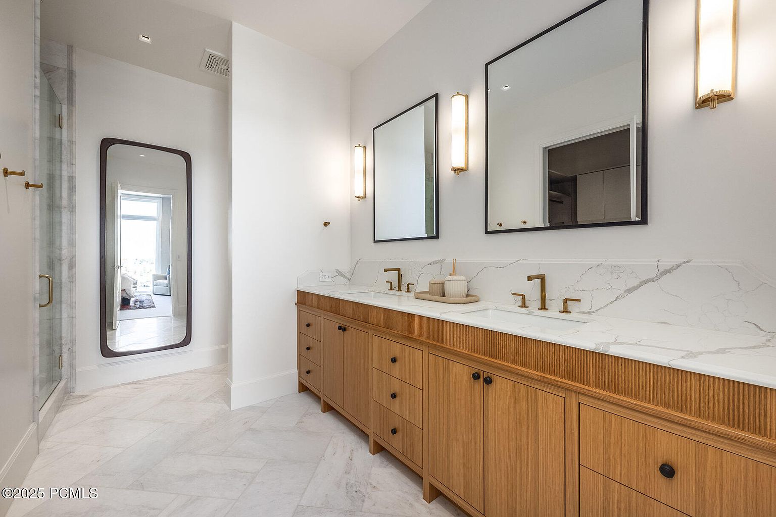 This is a primary bathroom featuring a double vanity with light wood cabinetry, a white marble countertop, and modern gold fixtures. Two rectangular mirrors hang above the sinks, illuminated by vertical sconces. The floor is covered in marble tiles, and a glass-enclosed shower is visible to the left, creating a luxurious and spa-like atmosphere.