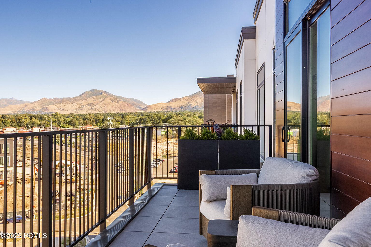 This image showcases a modern balcony with comfortable outdoor seating, including cushioned chairs and a small side table. The balcony features a sleek black railing and overlooks a scenic view of mountains and a developing landscape. The overall impression is one of relaxation and contemporary style, perfect for enjoying the outdoors.