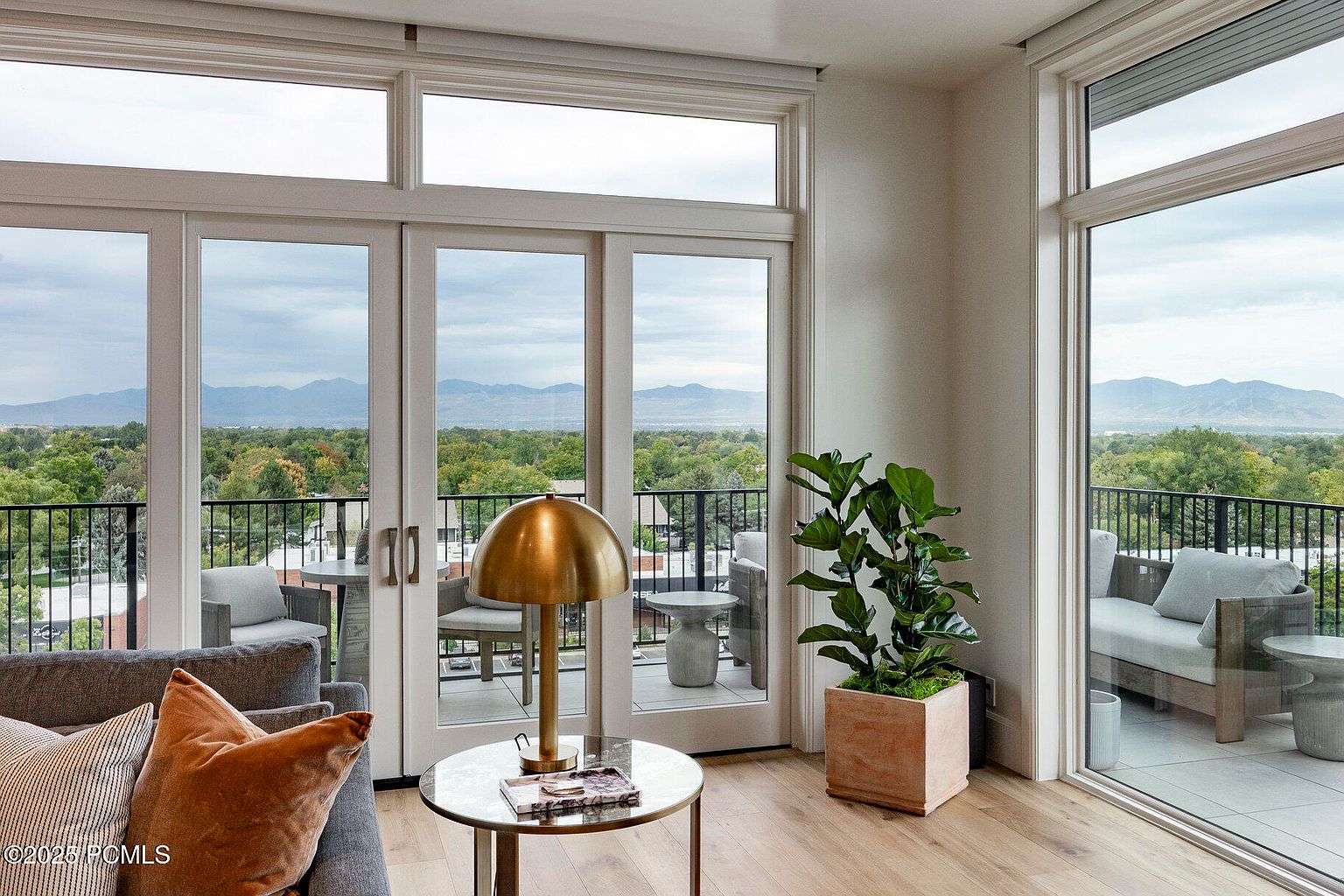 This is an interior shot of a living room, showcasing a bright and airy space with large windows offering a scenic view of mountains and greenery. The room features modern furniture, including a gray sofa with decorative pillows, a round side table with a gold lamp, and a potted plant, creating a comfortable and stylish atmosphere. The balcony is visible through the glass doors, extending the living space outdoors.