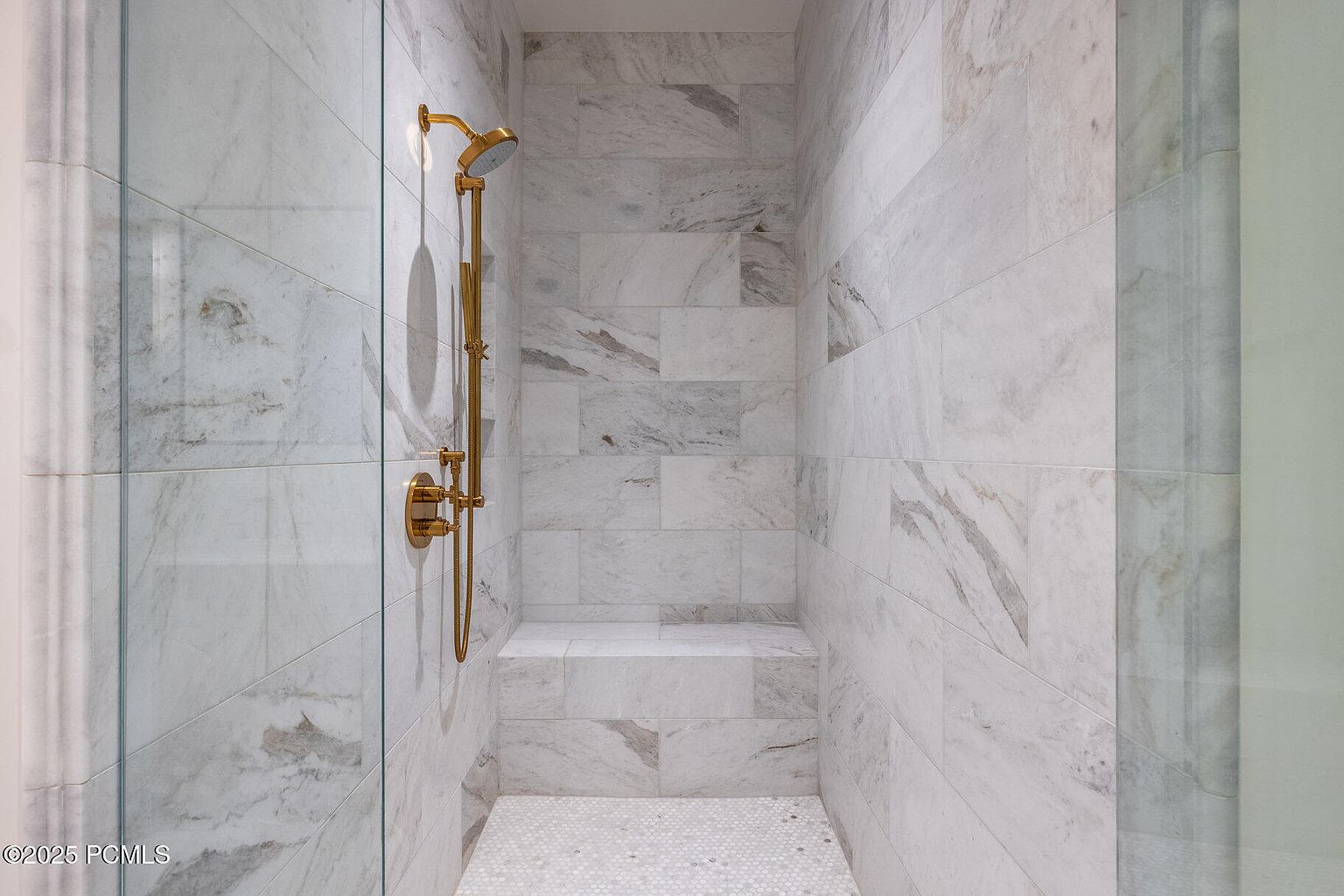 This is a well-lit primary bathroom showcasing a luxurious shower. The shower features marble tiling on the walls and a mosaic tile floor, complemented by gold fixtures. A built-in bench provides seating, enhancing the spa-like atmosphere.