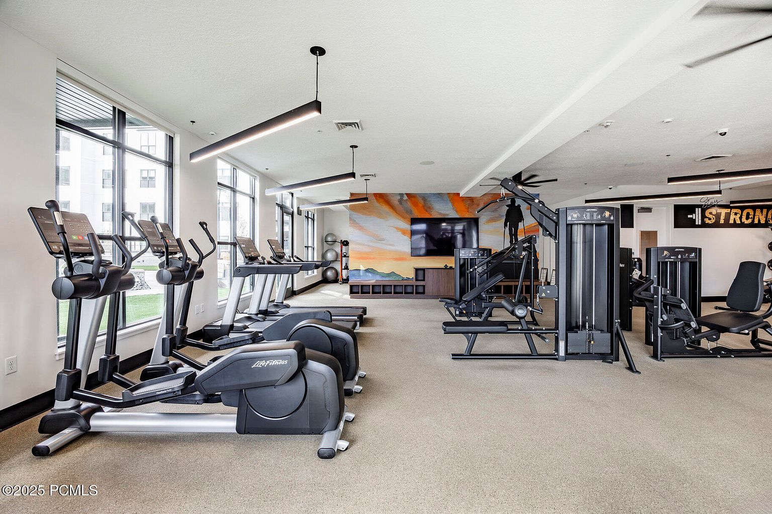 This is an interior shot of a well-equipped home gym. The space features a variety of exercise machines, including treadmills, elliptical trainers, and weightlifting equipment. Large windows provide natural light, and the walls are decorated with motivational artwork. The overall impression is a clean and modern fitness environment.