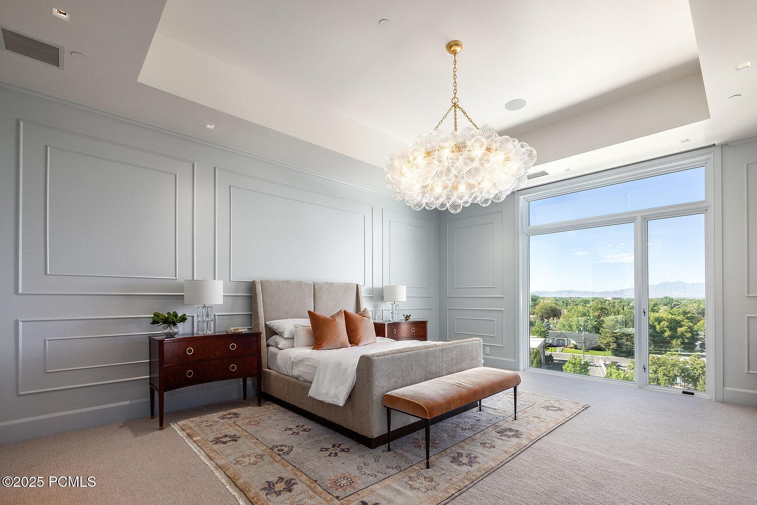 This is a luxurious primary bedroom featuring a large bed with a neutral-toned upholstered headboard, complemented by a pair of dark wood nightstands and table lamps. The room is decorated with elegant wall paneling and a striking bubble chandelier, while a large window offers a view of the landscape. A plush rug anchors the space, adding warmth and texture.