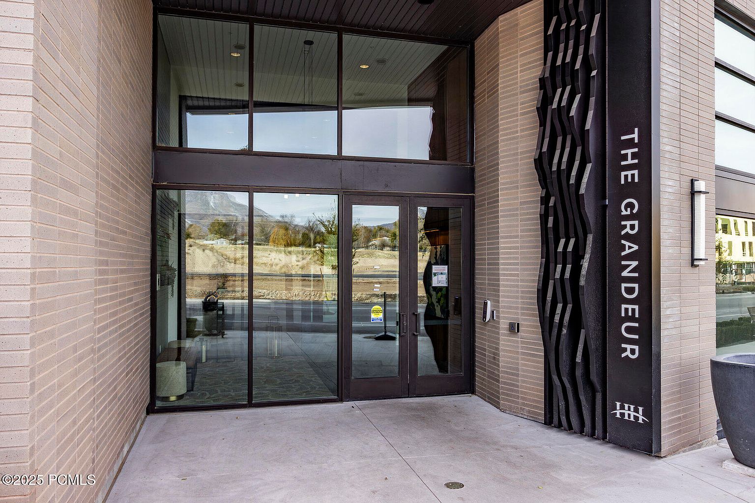 The image showcases the entryway of a building named "The Grandeur", featuring a modern architectural design with a brick facade and large glass windows and doors. A decorative black panel with vertical wave-like patterns adds visual interest to the entrance. The entryway is clean and well-maintained, suggesting a high-end residential or commercial property.