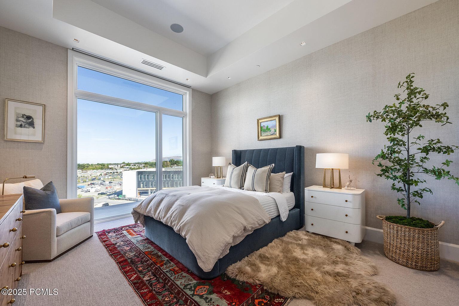 This is a primary bedroom featuring a large window with a view, a blue upholstered bed with neutral bedding, and matching white nightstands with lamps. The room is decorated in neutral tones with a textured wall covering, and an area rug adds warmth and color. A potted tree adds a touch of nature to the space.