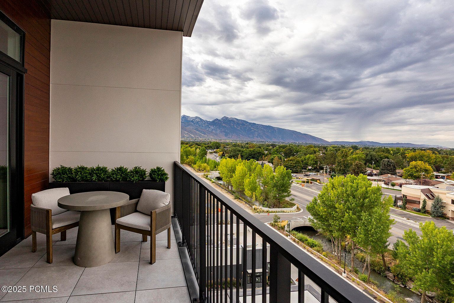 This image showcases a stylish balcony with a modern outdoor seating arrangement, including a round table and two chairs. The balcony features a sleek black railing and offers a scenic view of the surrounding landscape, including trees, a road, and distant mountains under a cloudy sky. The overall impression is one of sophisticated outdoor living.