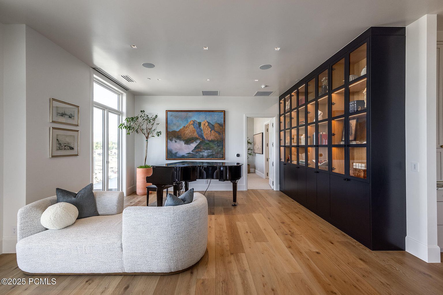 This is a bright and spacious living room featuring hardwood floors, a modern curved sofa with throw pillows, and a black grand piano beneath a landscape painting. A large built-in cabinet with glass doors displays decorative items, and natural light streams in through a large window, creating an inviting and elegant atmosphere. The room is designed with a blend of modern and classic elements.