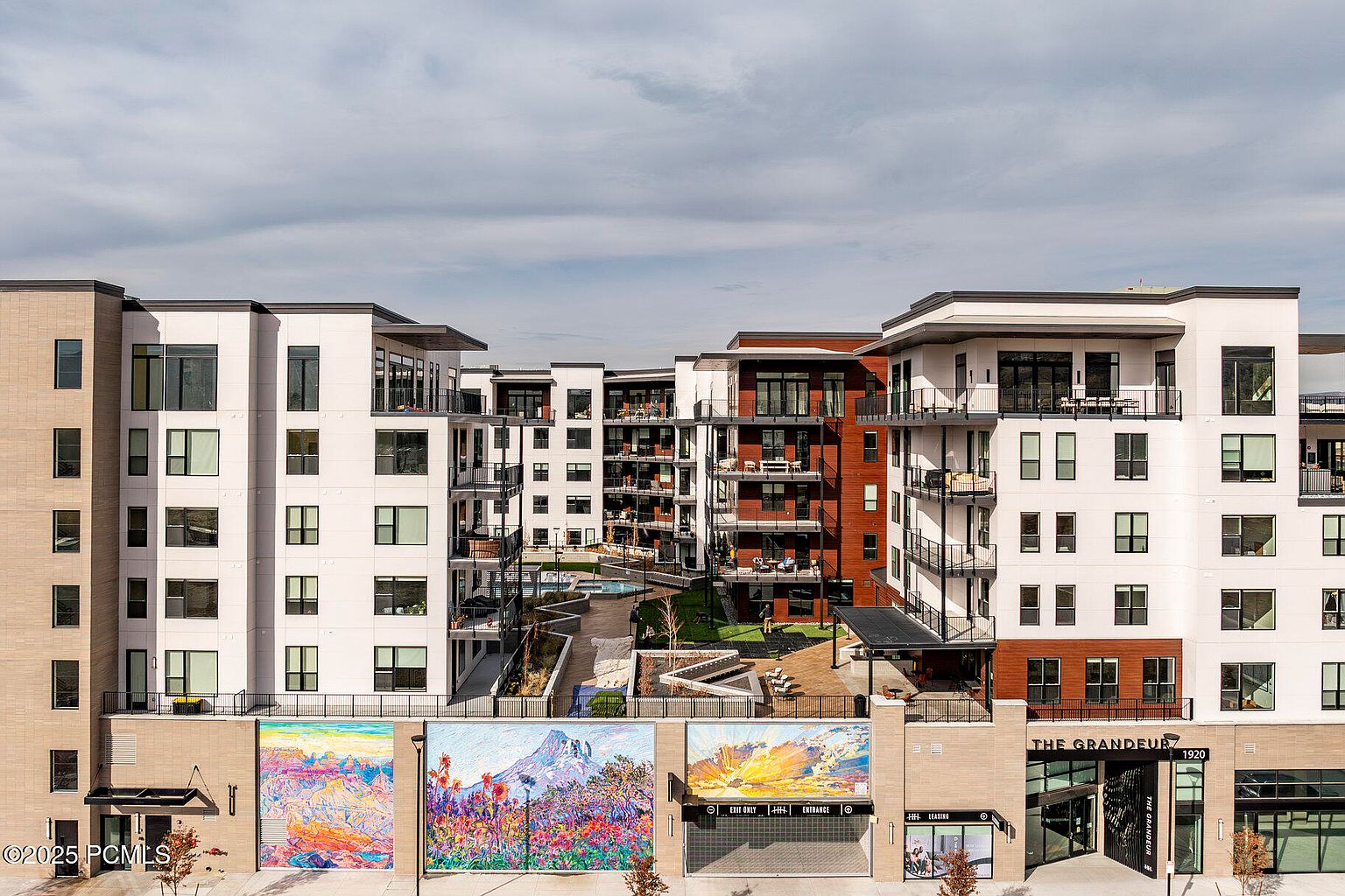 This image showcases the front view of a modern apartment complex. The buildings feature a mix of white and wood paneling, with multiple balconies and large windows. A brick facade at the base is adorned with colorful murals, and the entrance is clearly marked with the building's name, "The Grandeur," and address, "1920."