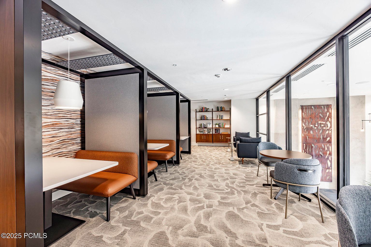 This interior shot showcases a modern office or study area with a mix of booth seating and individual tables, creating a versatile workspace. The design features a neutral color palette with pops of brown and gray, complemented by a patterned carpet and natural light streaming through large windows. A bookshelf adds a touch of sophistication and functionality to the space.