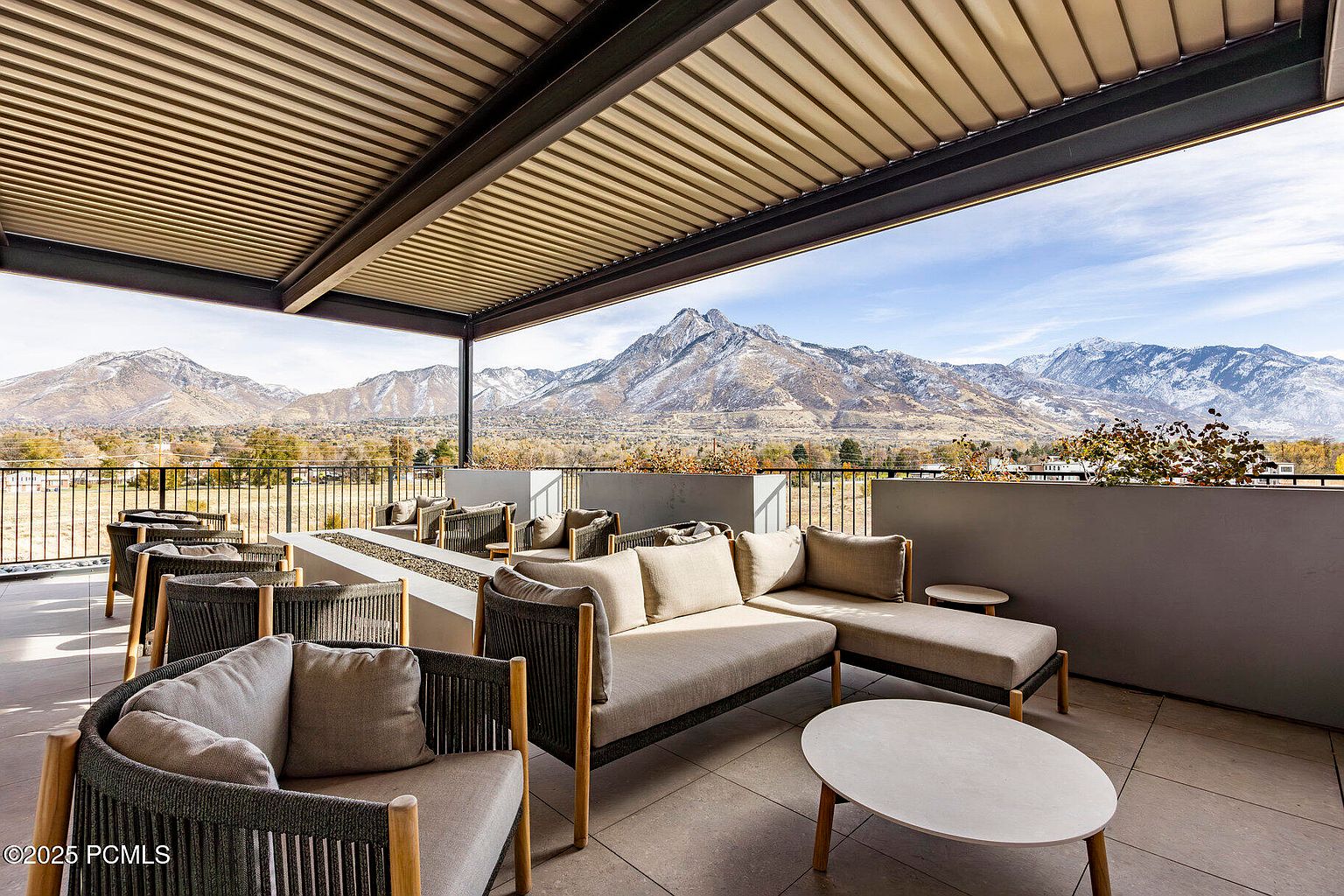 This image showcases a luxurious outdoor patio or balcony area, furnished with comfortable seating arrangements and a modern fire pit. The space is covered by a sleek, slatted roof structure, providing shade and architectural interest. The backdrop features a stunning mountain view, enhancing the appeal of this outdoor living space.