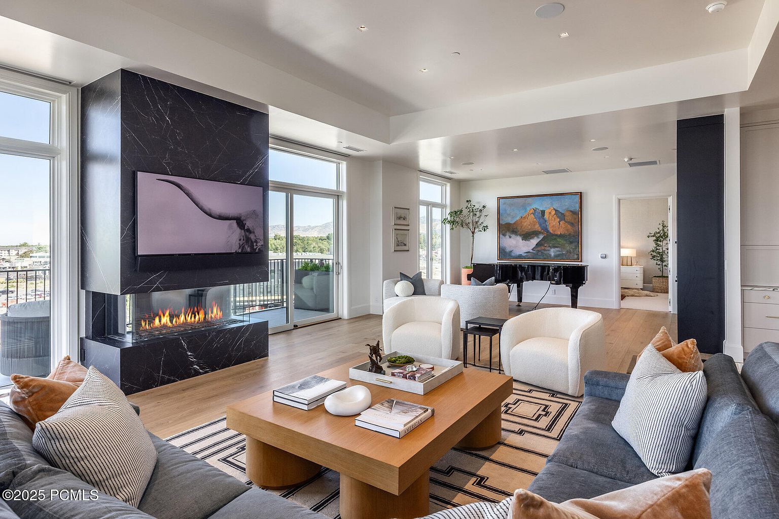 This is a luxurious living room featuring a modern fireplace with a television mounted above, surrounded by floor-to-ceiling windows offering scenic views. The room is furnished with comfortable sofas, armchairs, and a stylish coffee table, complemented by a grand piano in the background. The overall aesthetic is elegant and sophisticated, creating a welcoming and upscale atmosphere.