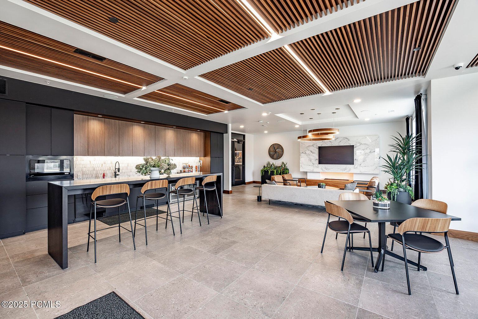 This image showcases a modern clubhouse amenity space, featuring a stylish kitchen area with dark cabinetry and bar seating, seamlessly transitioning into a comfortable lounge area with a fireplace and contemporary furnishings. The space is well-lit with a combination of natural and artificial light, highlighting the sleek design and inviting atmosphere, perfect for resident gatherings and relaxation.