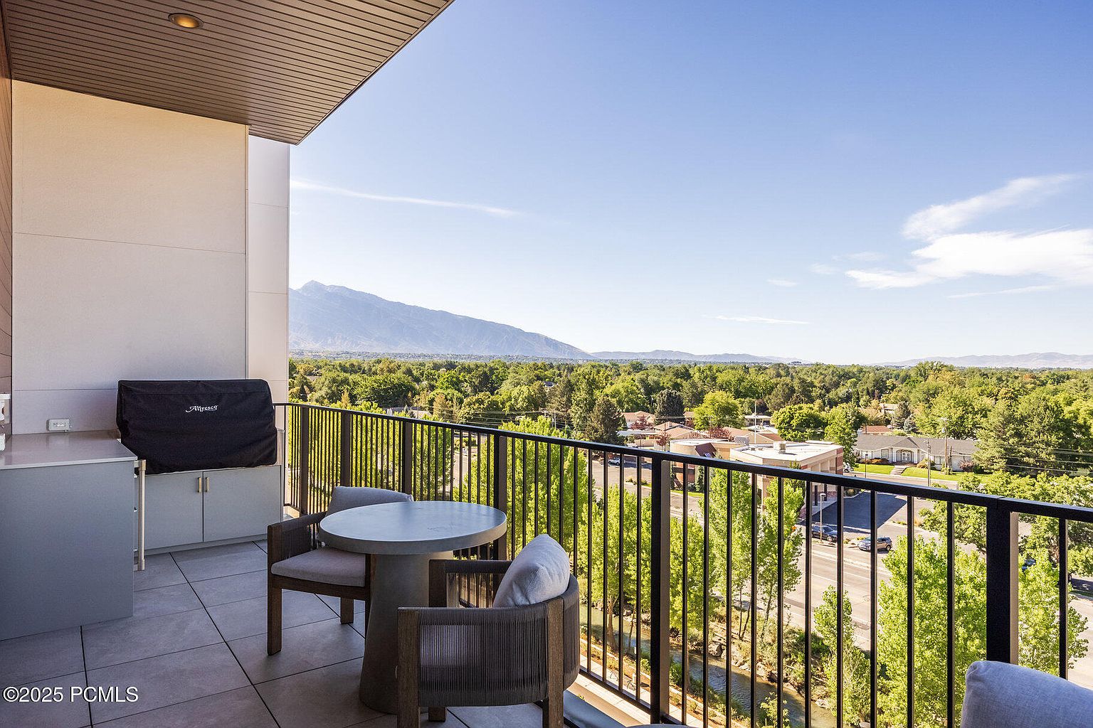 This image showcases a spacious balcony with a stunning view of the surrounding landscape. The balcony features a dining set with a round table and comfortable chairs, perfect for enjoying meals outdoors. A built-in grill area adds to the functionality of the space, making it ideal for entertaining. The black metal railing provides safety without obstructing the scenic vista of trees, mountains, and a clear blue sky.