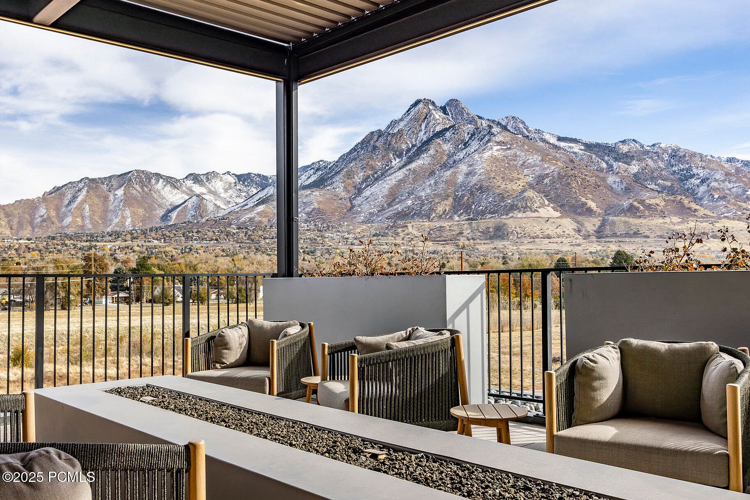 This image showcases a luxurious outdoor patio or balcony area with comfortable seating arrangements around a modern fire pit. The space is designed with a contemporary aesthetic, featuring sleek furniture and a minimalist color palette. The backdrop reveals a stunning mountain view, enhancing the property's appeal and offering a serene outdoor living experience.