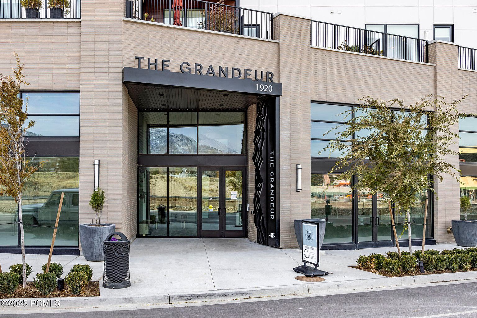 The image showcases the entryway of "The Grandeur" building, featuring a modern architectural design with a brick facade and a prominent black awning. Large glass windows and doors provide ample natural light and a view into the interior. Landscaping elements, including trees and shrubs, add to the building's curb appeal, while a sign and trash receptacle are placed near the entrance.