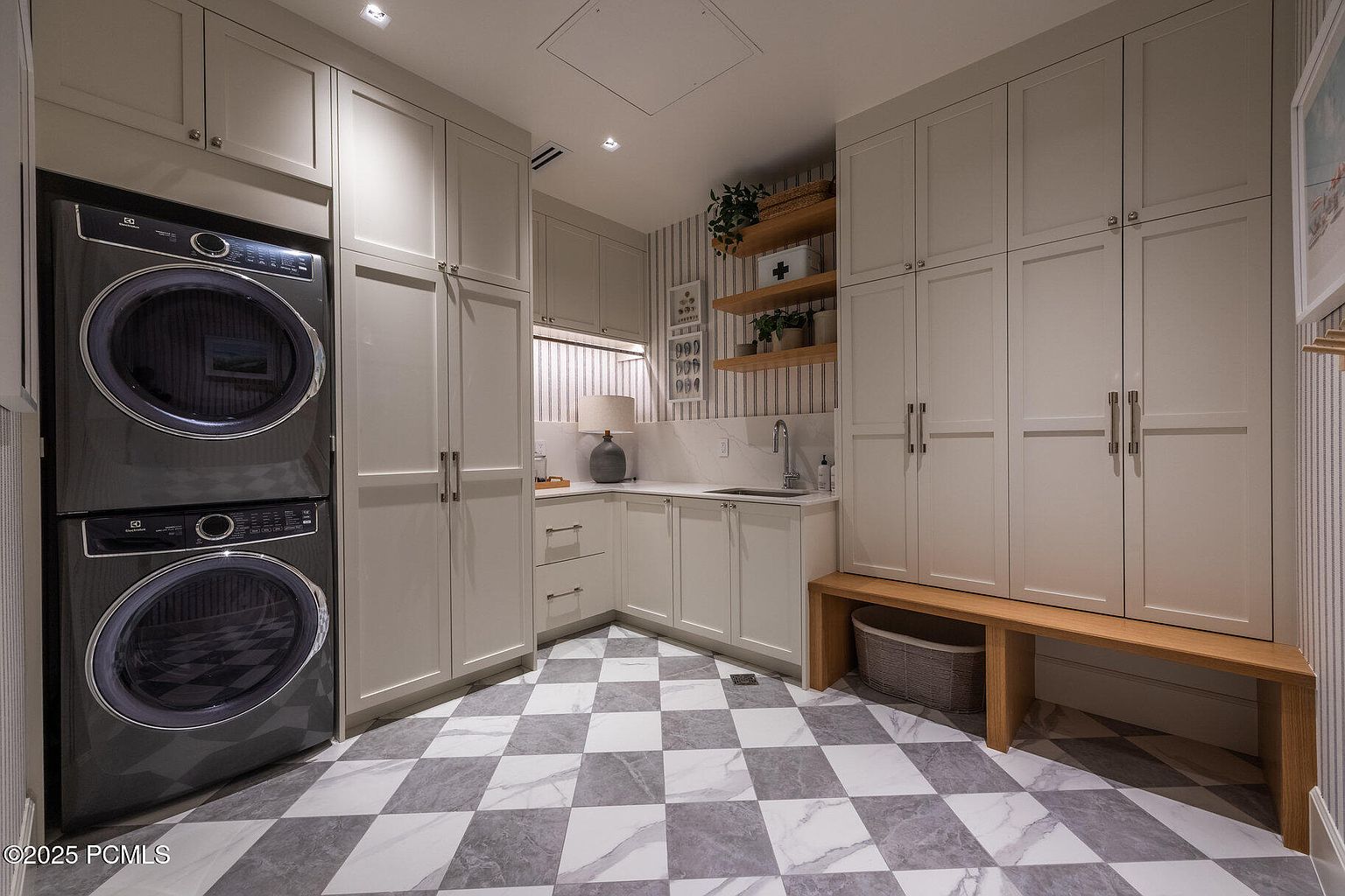 This is a well-organized laundry room featuring stacked washer and dryer units, ample storage cabinets, and a countertop with a sink. The room has a modern aesthetic with a checkered floor pattern and neutral color palette. A wooden bench provides a seating area, and open shelves add decorative elements.