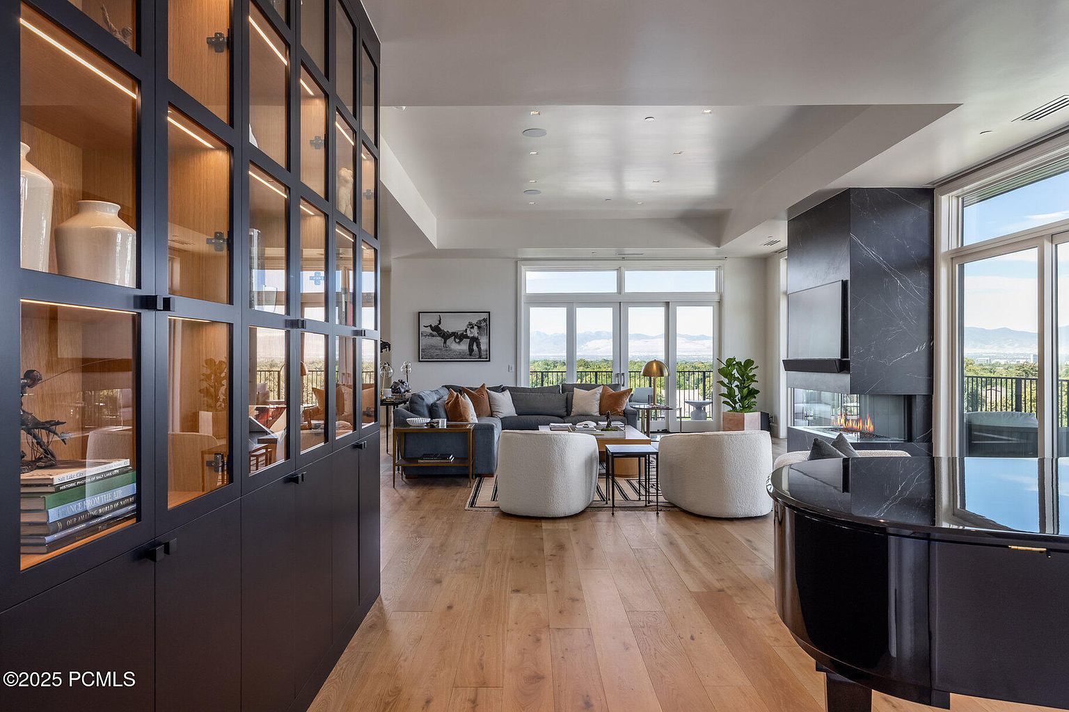 This is a spacious and luxurious living room featuring hardwood floors, a modern fireplace with a mounted TV, and large windows offering scenic views. The room is furnished with a comfortable sectional sofa, two round armchairs, and a grand piano, creating an elegant and inviting atmosphere. A large built-in cabinet with glass doors displays decorative items, adding to the room's sophisticated style.