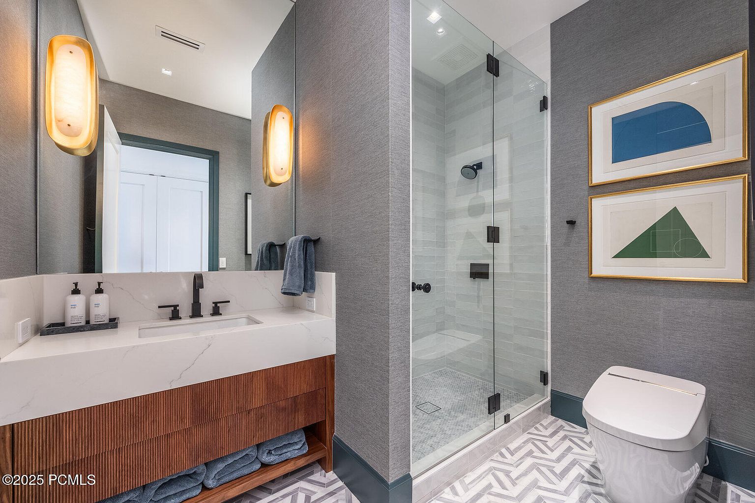This is a modern bathroom featuring a white marble countertop with a dark faucet and a wooden vanity with open shelving. A glass-enclosed shower is visible to the right, and the walls are covered in a textured gray wallpaper. Two framed geometric artworks hang above the toilet, adding a touch of sophistication.