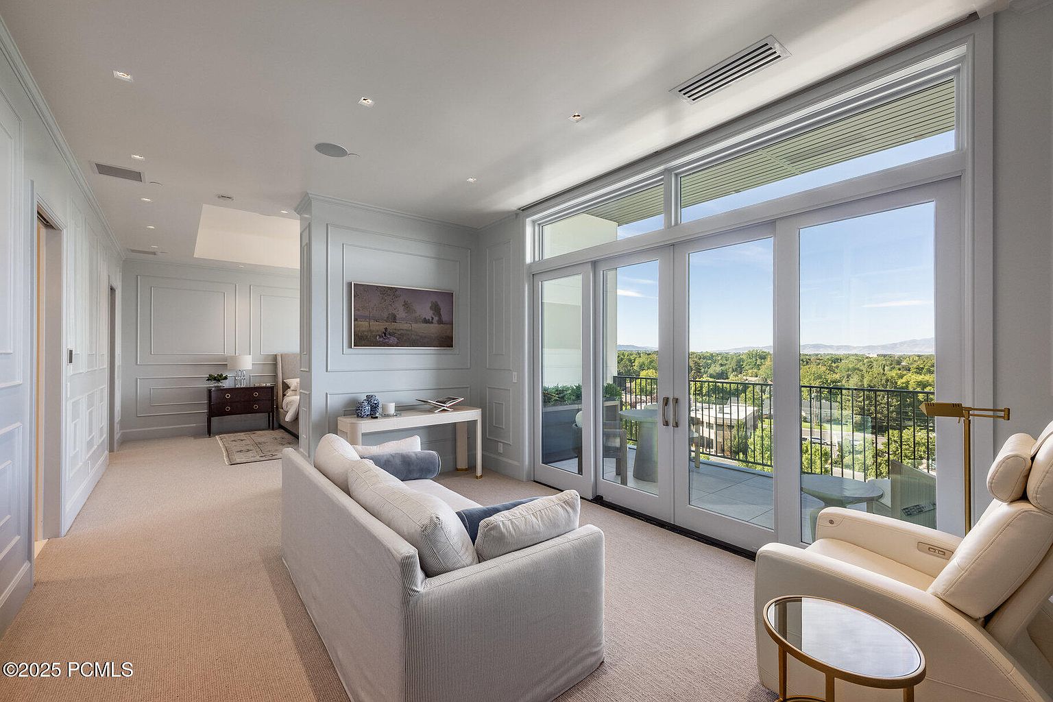 This is an interior shot of a living room featuring a white sofa, a white armchair, and a glass-topped side table. The room has large sliding glass doors that lead to a balcony with views of trees and mountains in the distance. The walls are light blue with white trim, and there is a painting on the wall above a console table.