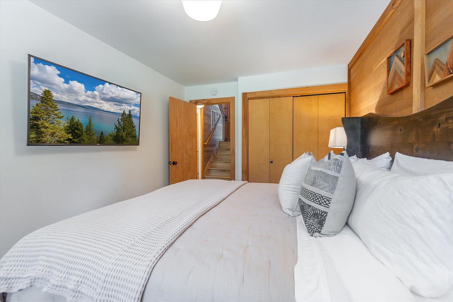 This cozy bedroom features a rustic aesthetic with a prominent wood-paneled accent wall behind the bed and a custom wood headboard. A large television is mounted on the wall, displaying a scenic landscape, while a doorway leads to a staircase visible in the background. The room is furnished with a plush bed dressed in neutral linens and decorative pillows, creating a warm and inviting atmosphere.