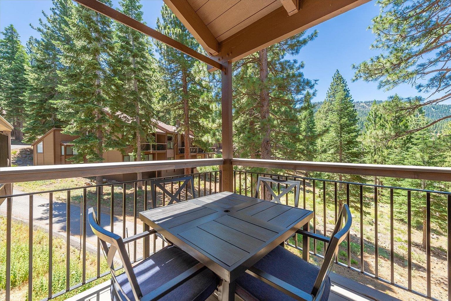 This elevated balcony features a dark metal railing and a square outdoor dining table with matching chairs, providing a perfect spot for al fresco dining. The space overlooks a lush, wooded landscape filled with tall pine trees and a glimpse of a neighboring building. The perspective is from the balcony looking outward, capturing a serene and private mountain-style setting.
