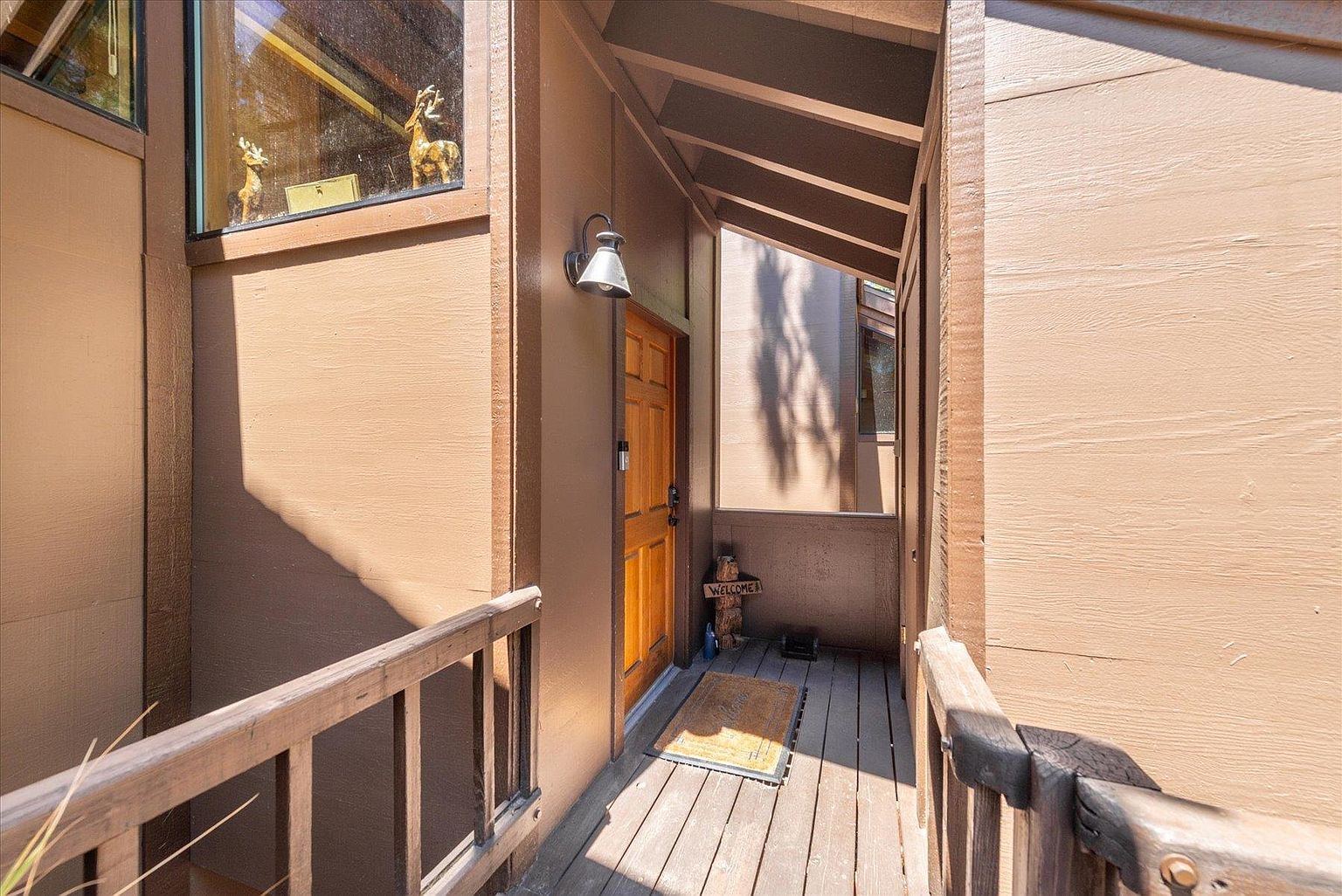 This entryway features a wooden deck leading to a warm-toned wooden front door, sheltered by an overhanging roof structure. The exterior walls are finished in a smooth, brown-painted material, complemented by a wall-mounted light fixture and a small 'Welcome' sign near the entrance. A wooden railing borders the deck, and large windows above reveal a glimpse of decorative deer figurines inside.