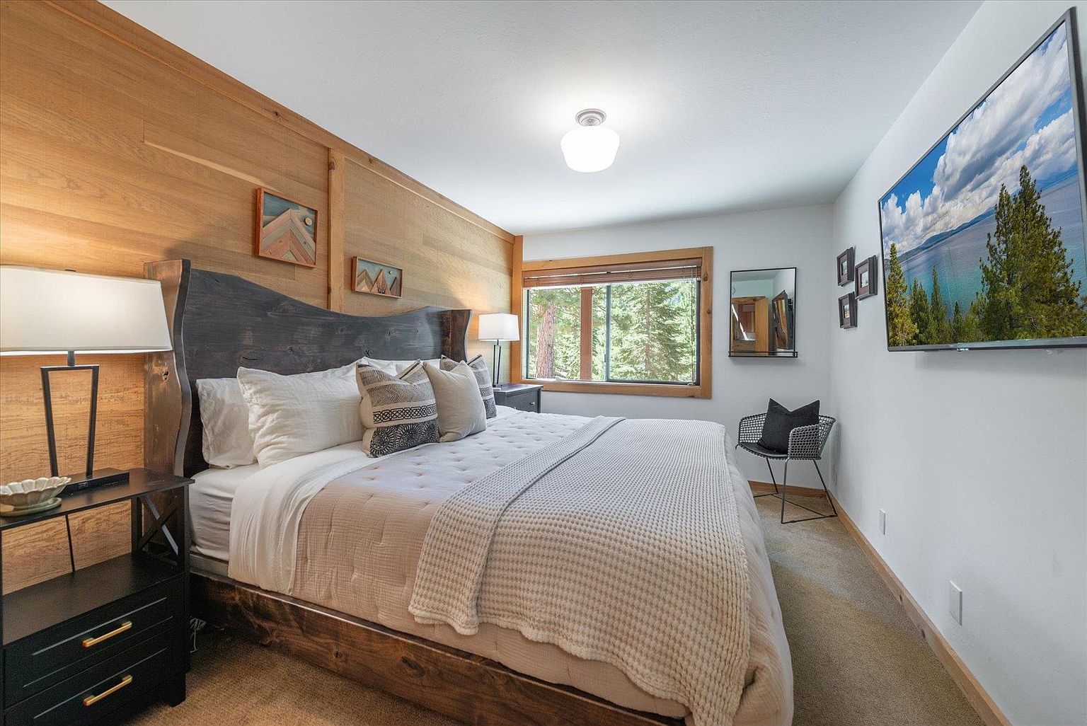 This cozy primary bedroom features a rustic aesthetic with a prominent wood-paneled accent wall behind a dark, live-edge wooden headboard. The room is furnished with a plush bed, a matching dark wood nightstand, and a modern wire-frame chair near a window that looks out onto a wooded area. A large flat-screen television is mounted on the wall, completing this comfortable and inviting mountain-style retreat.