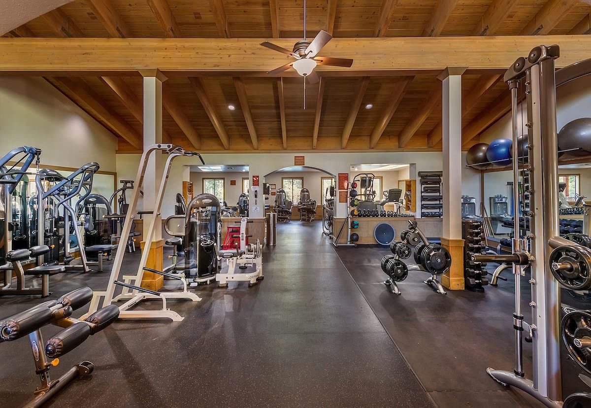 This spacious home gym features a high, vaulted ceiling with exposed wooden beams and a central ceiling fan. The room is equipped with a variety of professional-grade strength training machines, free weights, and exercise balls, all set against a dark, durable rubberized floor. The layout is open and well-lit, providing a functional and inviting space for fitness activities.