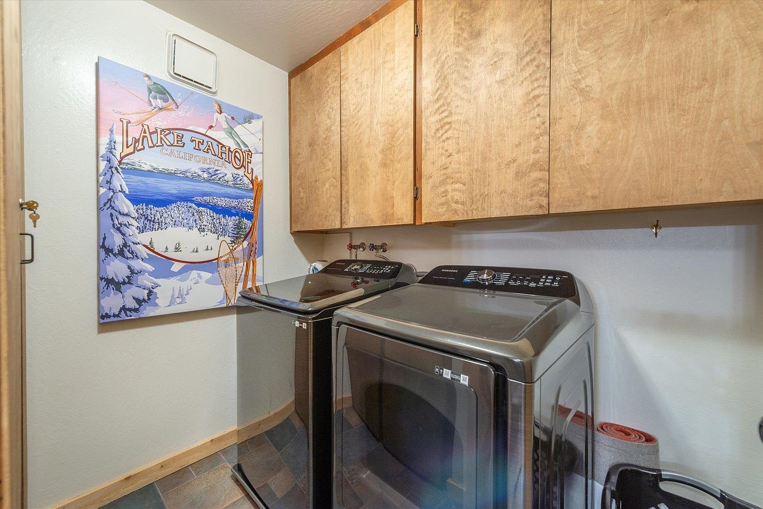 This functional laundry room features a side-by-side washer and dryer set in a metallic finish, positioned beneath light-toned wood cabinetry that provides ample storage. A large, decorative Lake Tahoe-themed wall art piece adds a touch of local character to the space, which is finished with neutral walls and dark, multi-toned floor tiles. The perspective is a straight-on shot, highlighting the efficient use of space in this utility area.