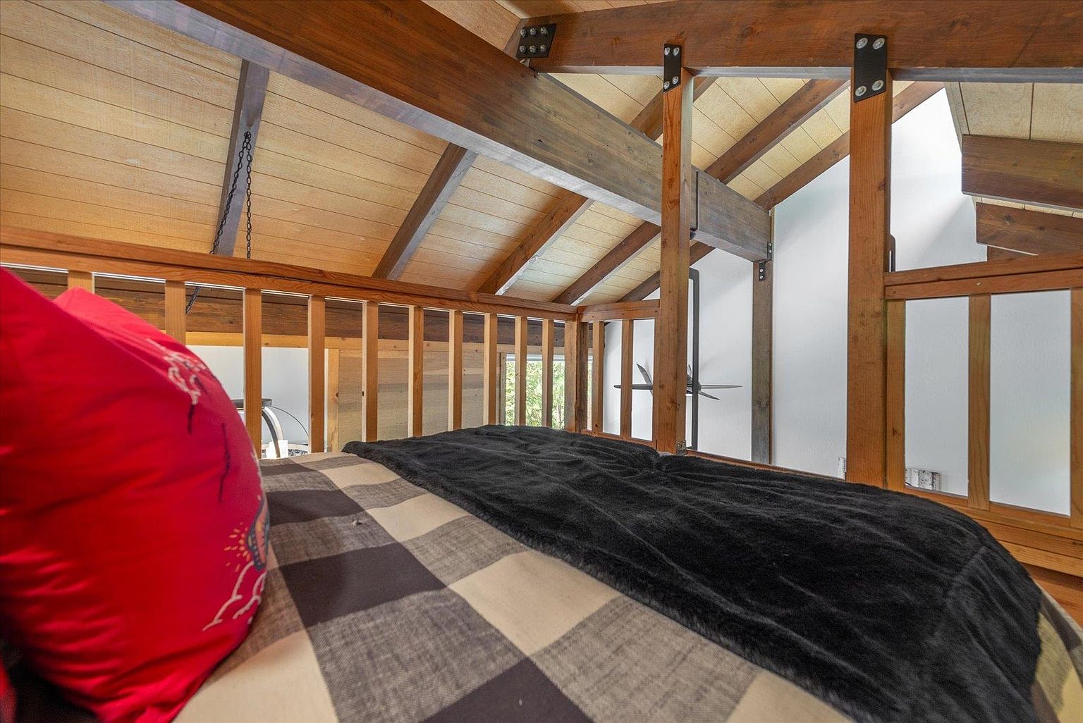 This image captures a cozy loft-style bedroom space, featuring a bed dressed in a plaid comforter and a bright red pillow. The area is framed by rustic exposed wooden beams and a wooden railing that overlooks the lower level of the home. The vaulted ceiling with light-toned wood paneling adds a warm, cabin-like aesthetic to this elevated sleeping nook.