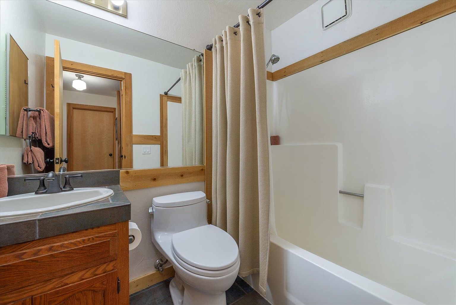 This compact bathroom features a vanity with a dark countertop, a white sink, and a wooden cabinet base. A large mirror spans the wall above the vanity, reflecting the adjacent doorway, while a beige shower curtain conceals the tub and shower combination. The room is accented with light-toned wood trim, creating a rustic and functional aesthetic.