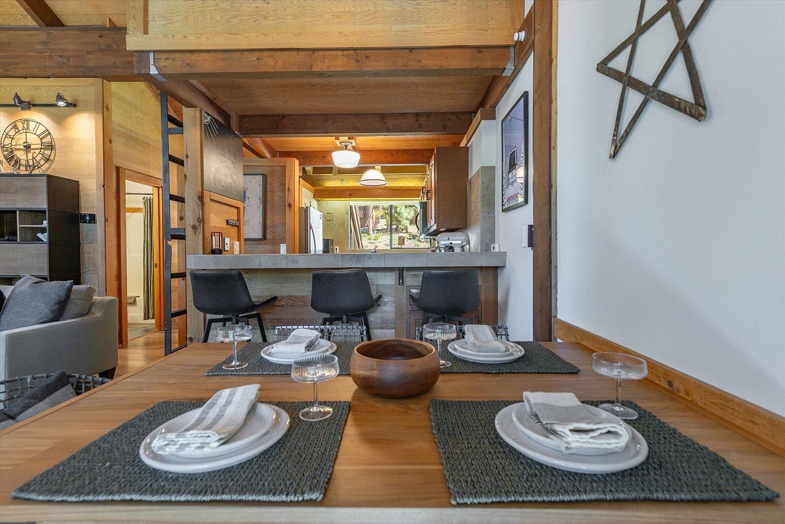 This dining area features a warm, rustic aesthetic with exposed wooden ceiling beams and a wooden dining table set for four. The space opens up to a kitchen breakfast bar with three dark stools, creating an inviting, open-concept layout. A decorative star wall hanging adds a touch of character to the neutral-toned walls, while the perspective looks directly from the dining table toward the kitchen area.