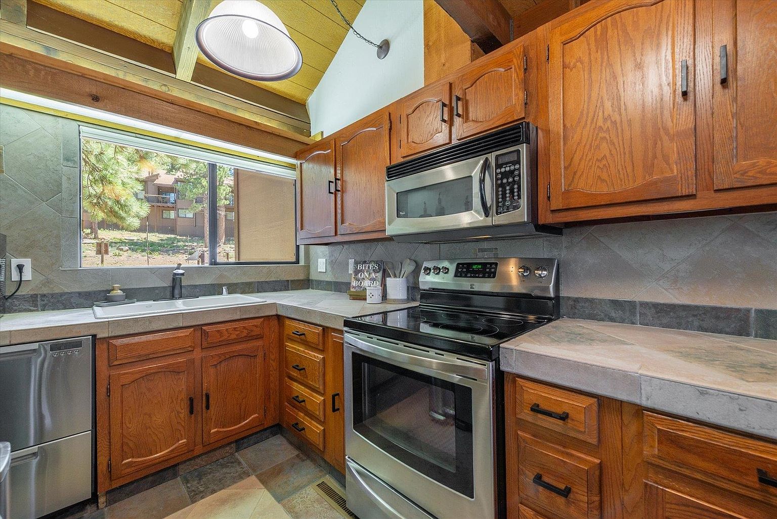 This kitchen features warm-toned wood cabinetry paired with neutral stone-look countertops and a tiled backsplash. The space is equipped with stainless steel appliances, including a microwave and a range, and is illuminated by a large pendant light hanging from a vaulted ceiling. A window above the sink provides a view of the outdoors, creating a bright and functional cooking area.