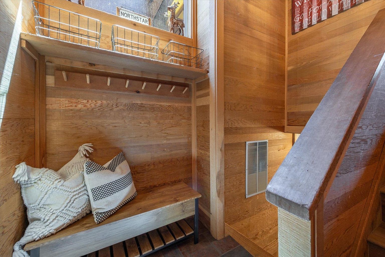 This cozy entryway features warm wood-paneled walls and a built-in bench with a lower storage shelf. Above the bench, a shelf with wire baskets provides additional organization, while a decorative sign and a small figurine add a welcoming touch. The perspective captures the transition from the entryway into a stairwell, highlighting the rustic, cabin-like aesthetic of the home.