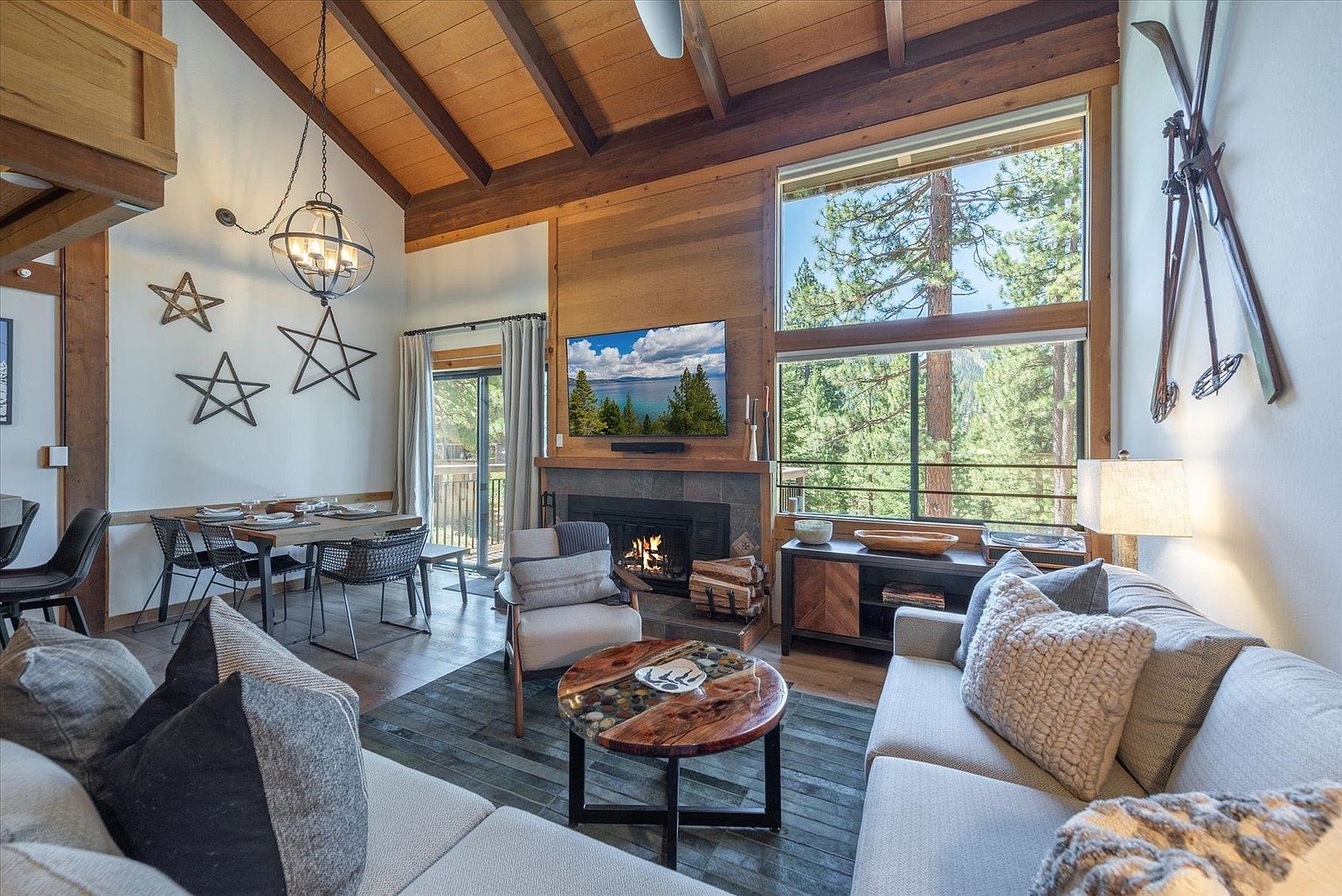 This open-concept living and dining area features a vaulted wood-paneled ceiling and a large window wall that frames a view of the surrounding forest. The living space is anchored by a stone-clad fireplace with a mounted television above, while a rustic wood-slab coffee table sits atop a textured area rug. The room is decorated with a cozy, mountain-inspired aesthetic, including vintage-style skis on the wall and star-shaped wall art, creating a warm and inviting atmosphere.