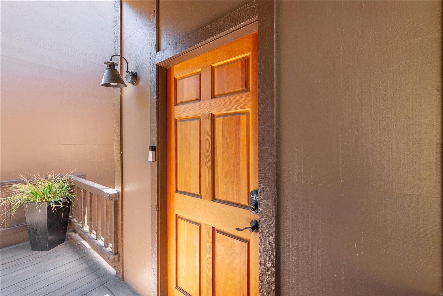 This image captures a private entryway featuring a warm-toned wooden door with a six-panel design and a modern electronic keypad lock. The entrance is situated on a wooden deck, accented by a potted plant and a classic wall-mounted lantern fixture. The perspective is a straight-on shot that highlights the inviting, rustic aesthetic of the home's exterior transition space.